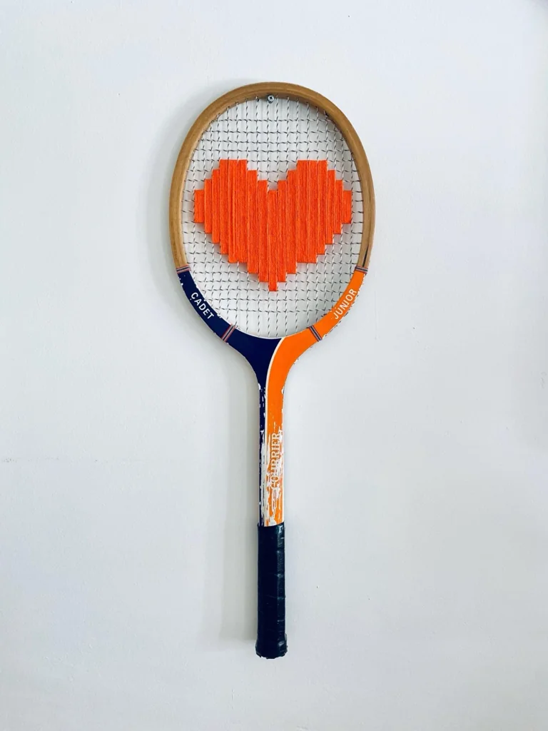 Raquette de tennis vintage en bois brodée d'un cœur pixel orange, accrochée sur un mur blanc