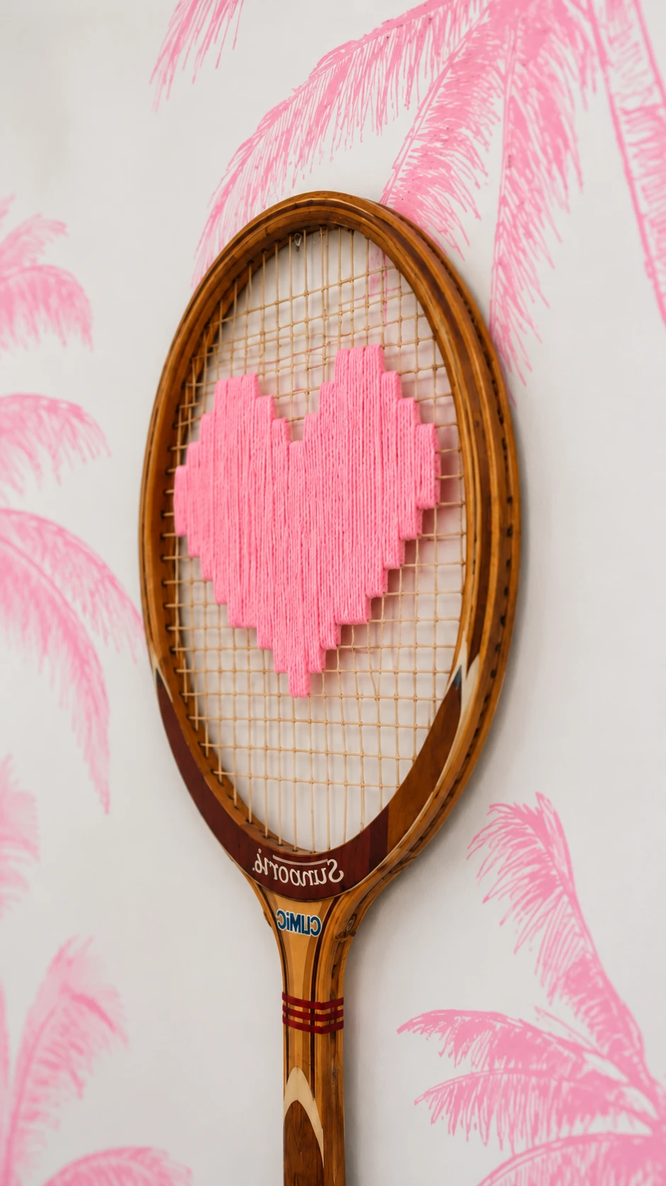 Gros plan d'une raquette vintage en bois au cœur rose brodé, sur un papier peint à palmiers roses