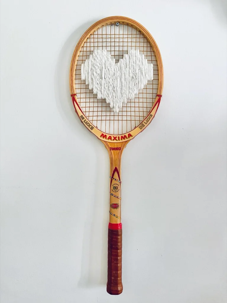 Raquette de tennis vintage en bois brodée d'un cœur pixel blanc velours, accrochée sur un mur blanc