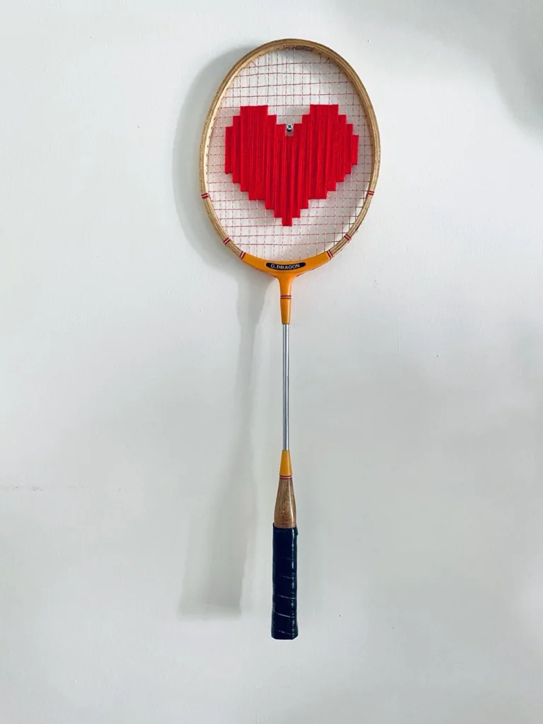 Raquette de badminton vintage en bois brodée d'un cœur pixel rouge, accrochée sur un mur blanc