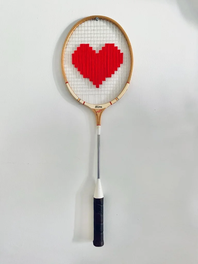 Raquette de badminton vintage en bois brodée d'un cœur pixel rouge, au col blanc et au manche noir, accrochée sur un mur blanc