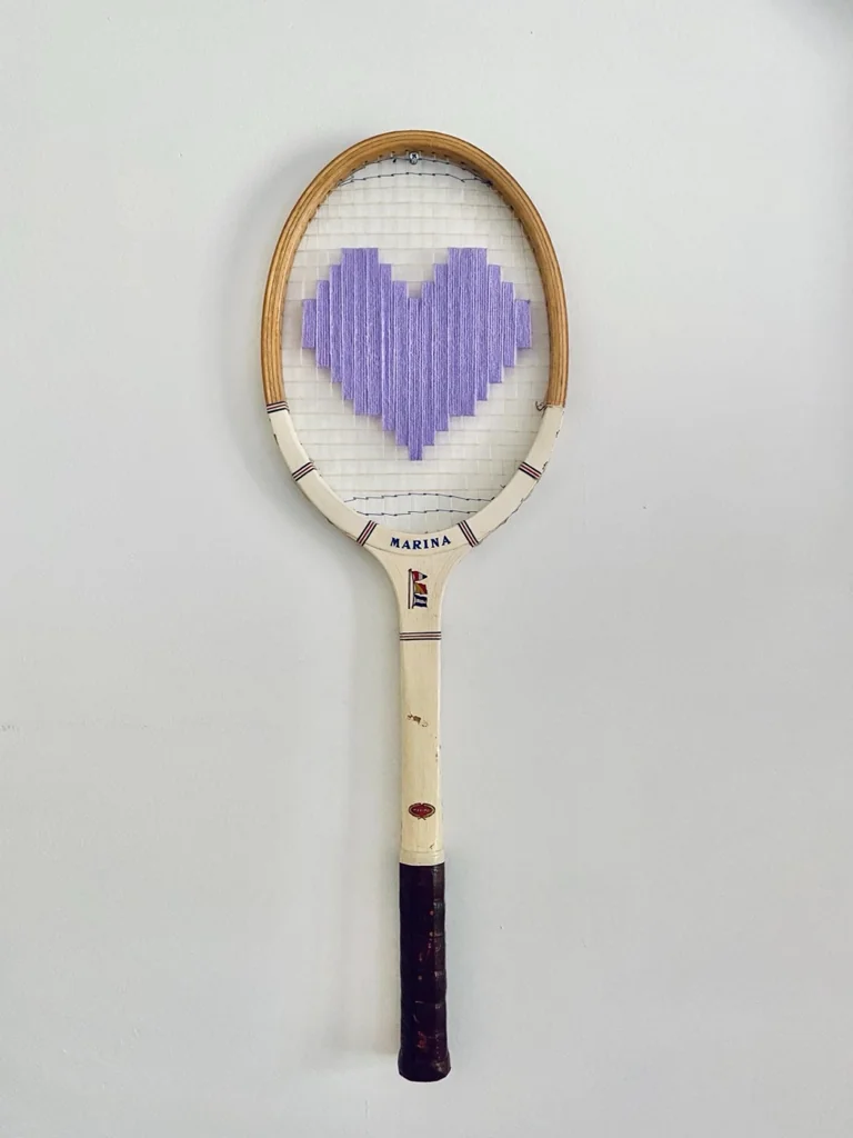 Raquette de tennis vintage en bois brodée d'un cœur pixel lavande, accrochée sur un mur blanc