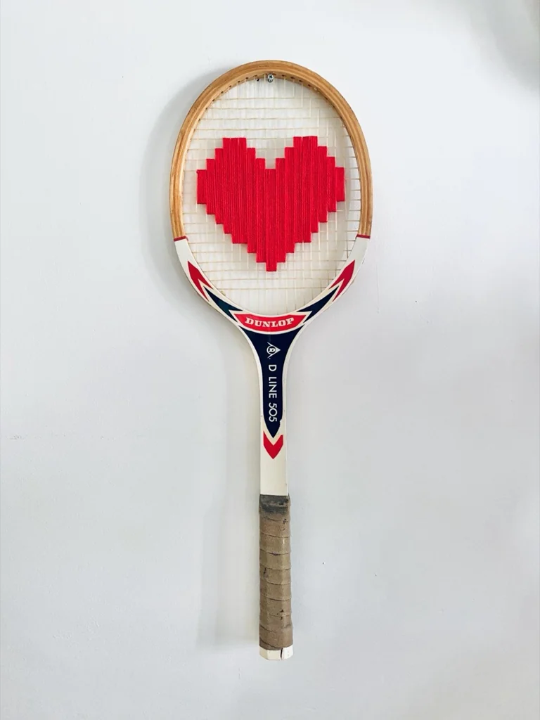 Raquette de tennis vintage en bois brodée d'un cœur pixel rouge, accrochée sur un mur blanc