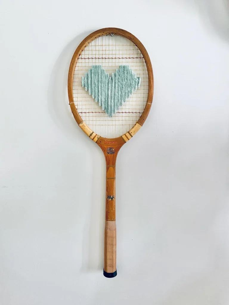 Raquette de tennis vintage en bois brodée d'un cœur pixel vert pastel, accrochée sur un mur blanc