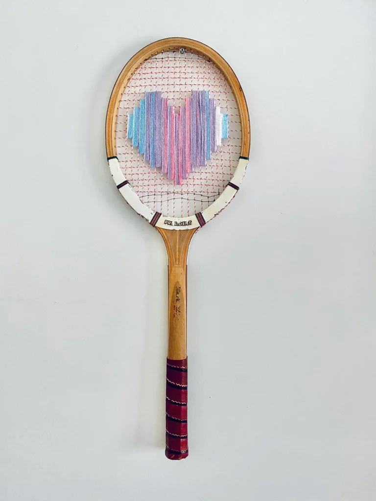 Raquette de tennis vintage en bois brodée d'un cœur pixel pastel multicolore, accrochée sur un mur blanc