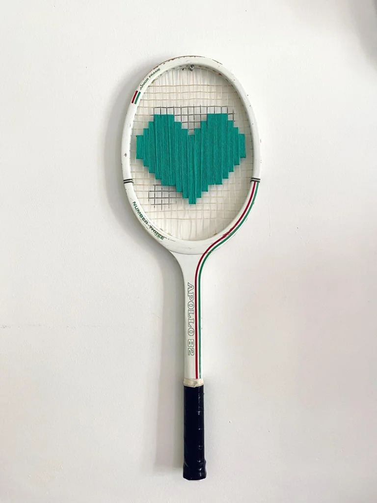 Raquette de tennis vintage en bois brodée d'un cœur pixel vert, accrochée sur un mur blanc