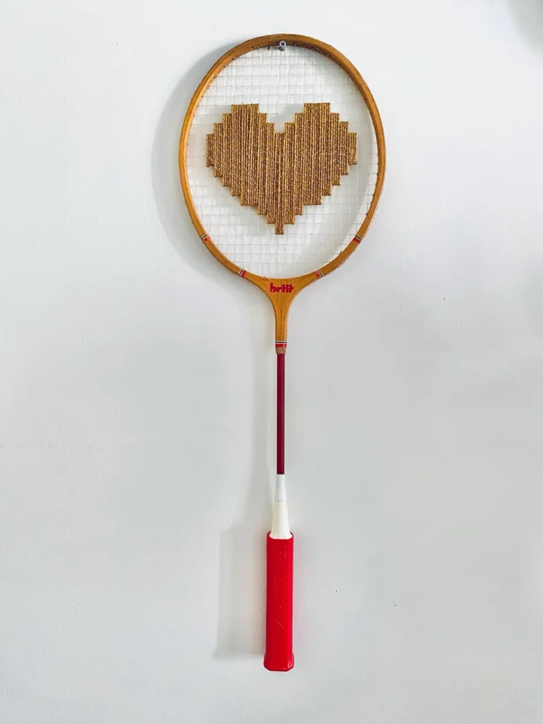 Raquette de badminton vintage en bois brodée d'un cœur pixel doré caramel, accrochée sur un mur blanc